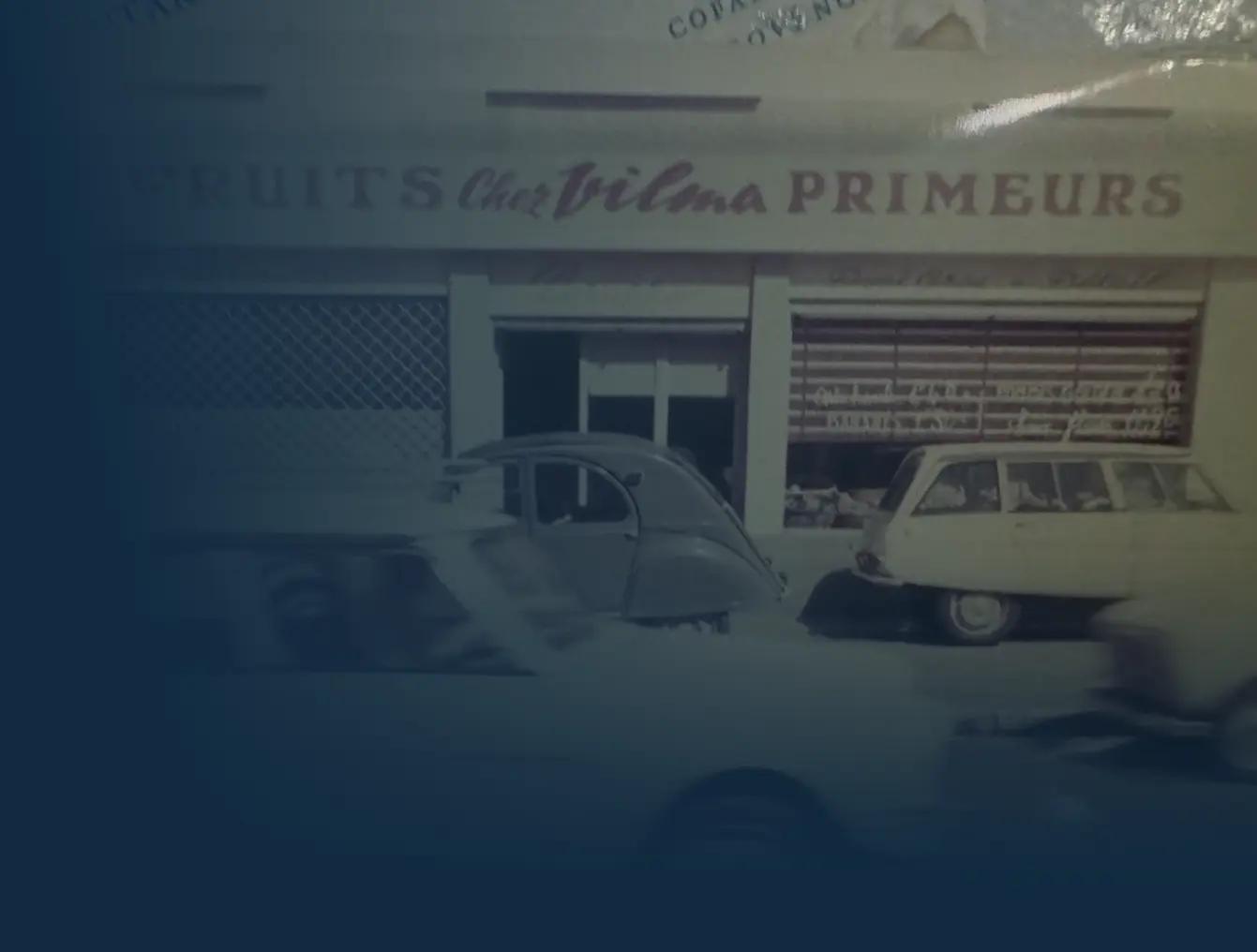 Des voitures anciennes garées devant un magasin de fruits aux enseignes défraîchies.